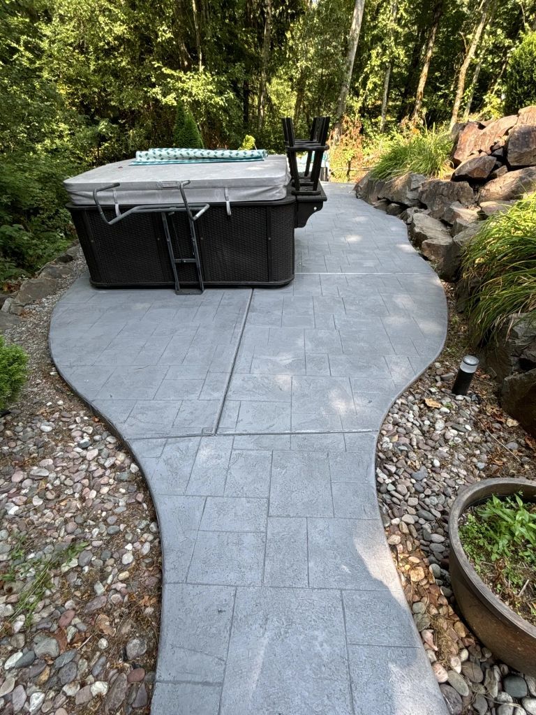 A hot tub sits on a gray stone pathway surrounded by landscaping and trees.