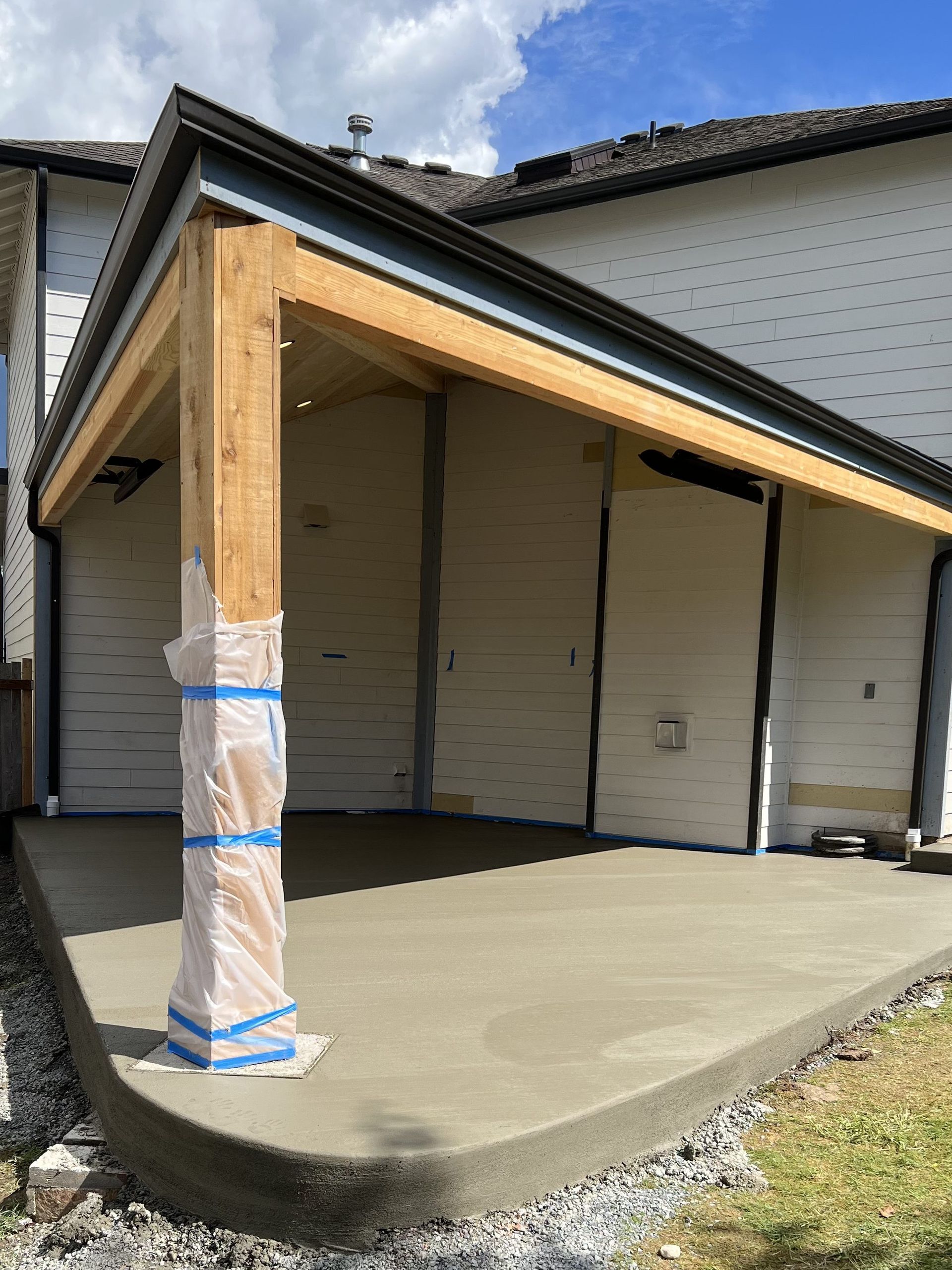 Newly poured concrete patio with a wooden beam porch roof attached to a white building. The porch support is wrapped in protective plastic.