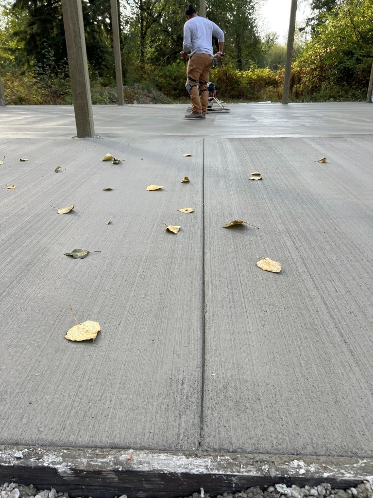 A worker smoothing a fresh concrete surface, with several fallen leaves scattered on the ground.