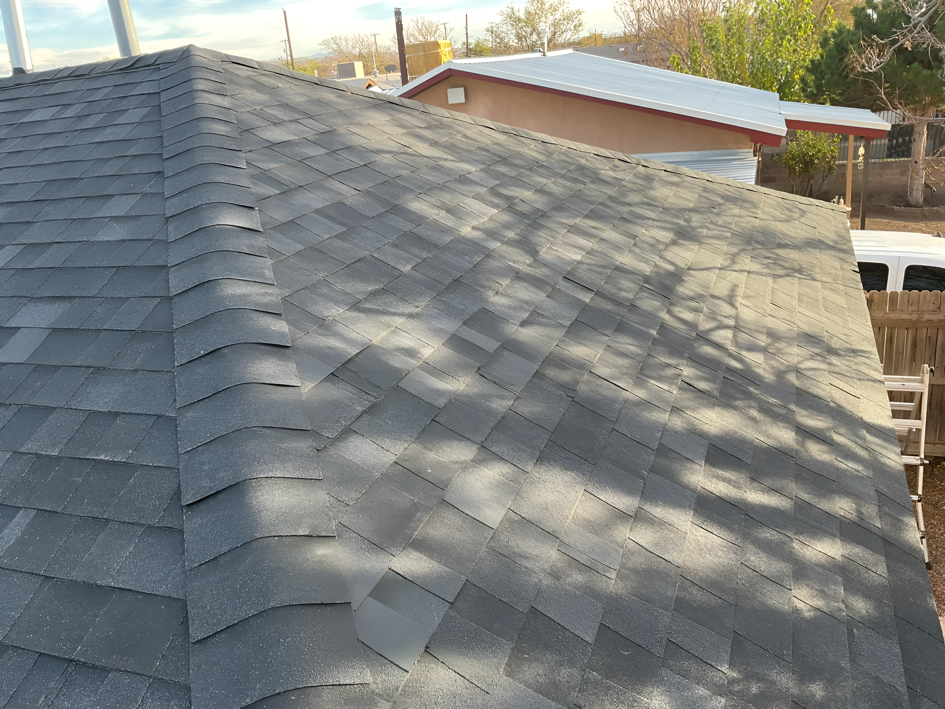 Gray asphalt shingle roof of a house with visible wear and tear, in an outdoor setting.