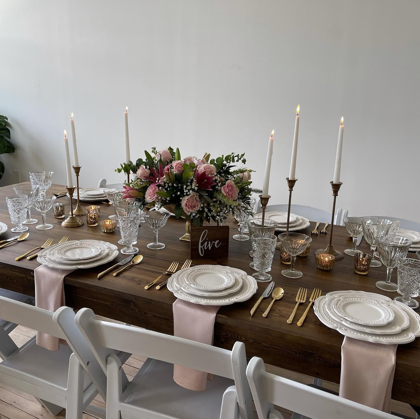 Formal dining table set with white dishes, gold cutlery, pink napkins, and a floral centerpiece.
