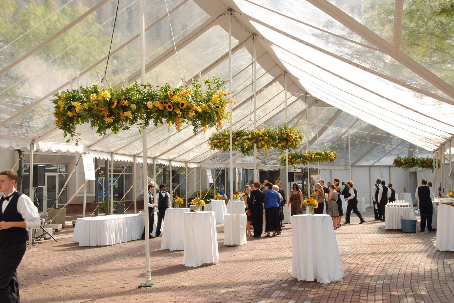 Clear tent with floral arrangements, cocktail tables, and people at an outdoor event.