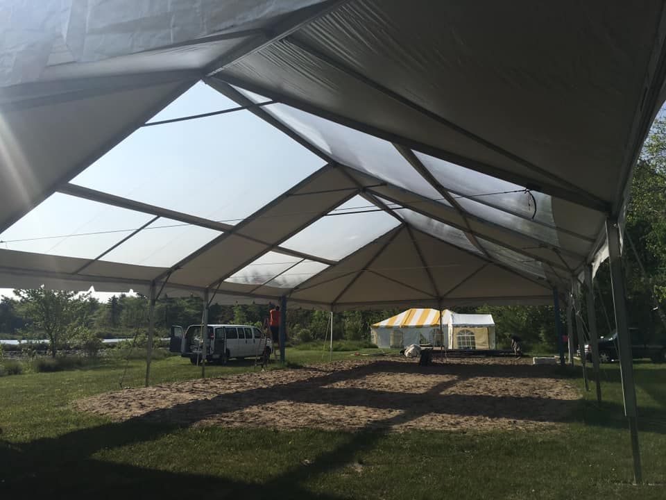Large tent with light tan roof over a grassy area. White vehicle and people visible inside.