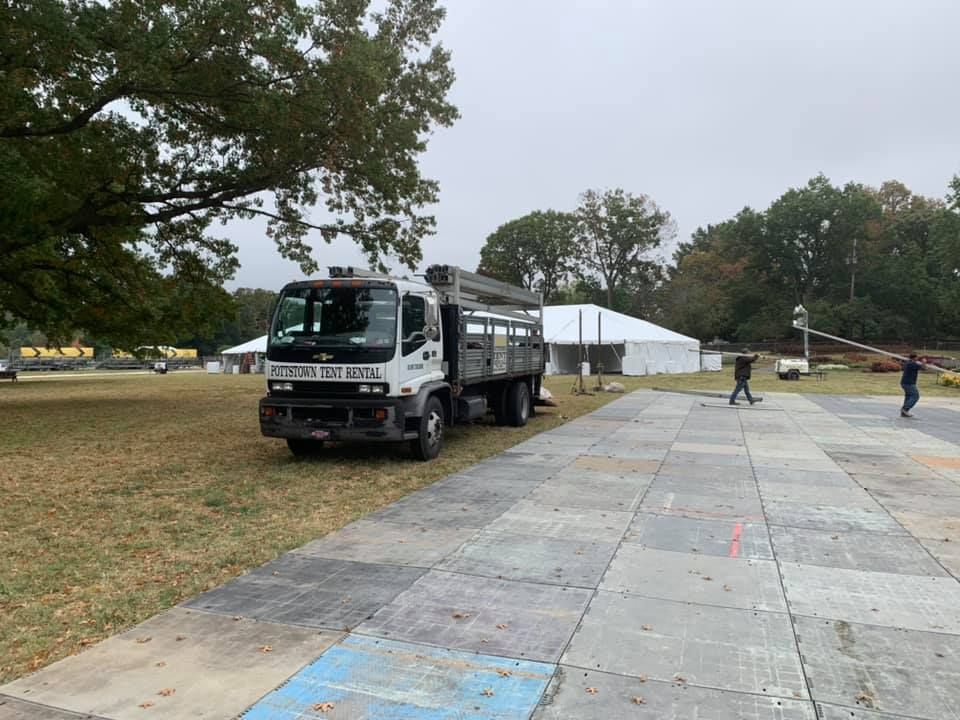 Truck parked near a large tent, two figures working, open grassy area, overcast sky.