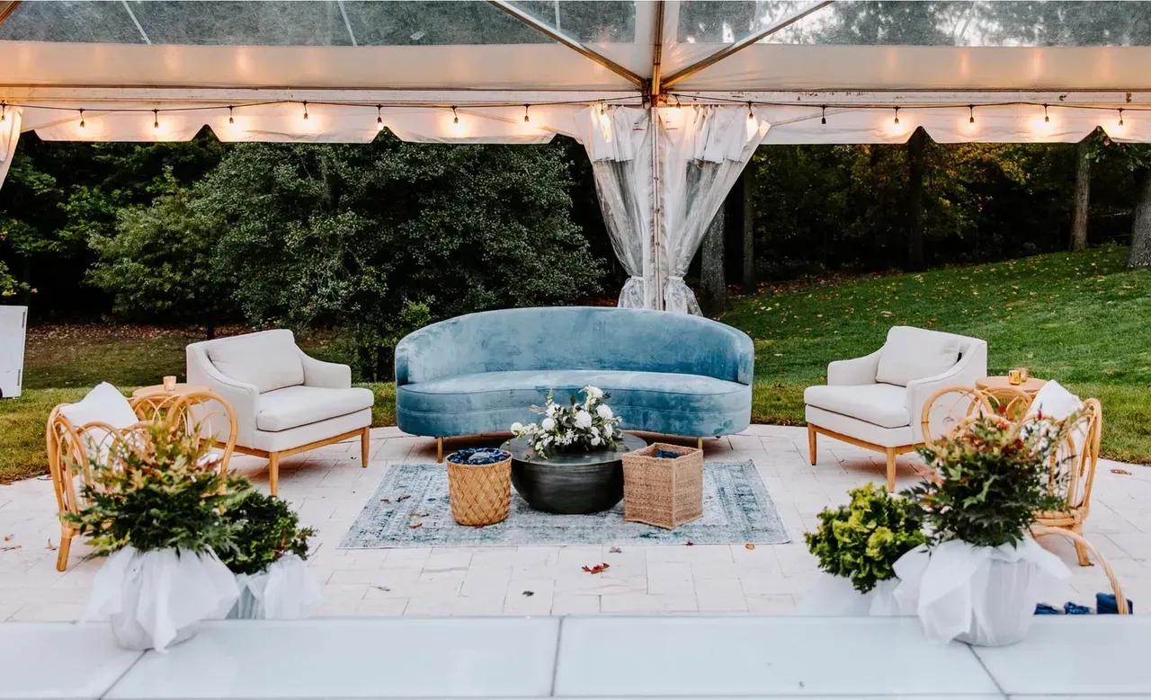 Outdoor lounge area with blue velvet sofa, wicker chairs, and small round tables under a clear tent.