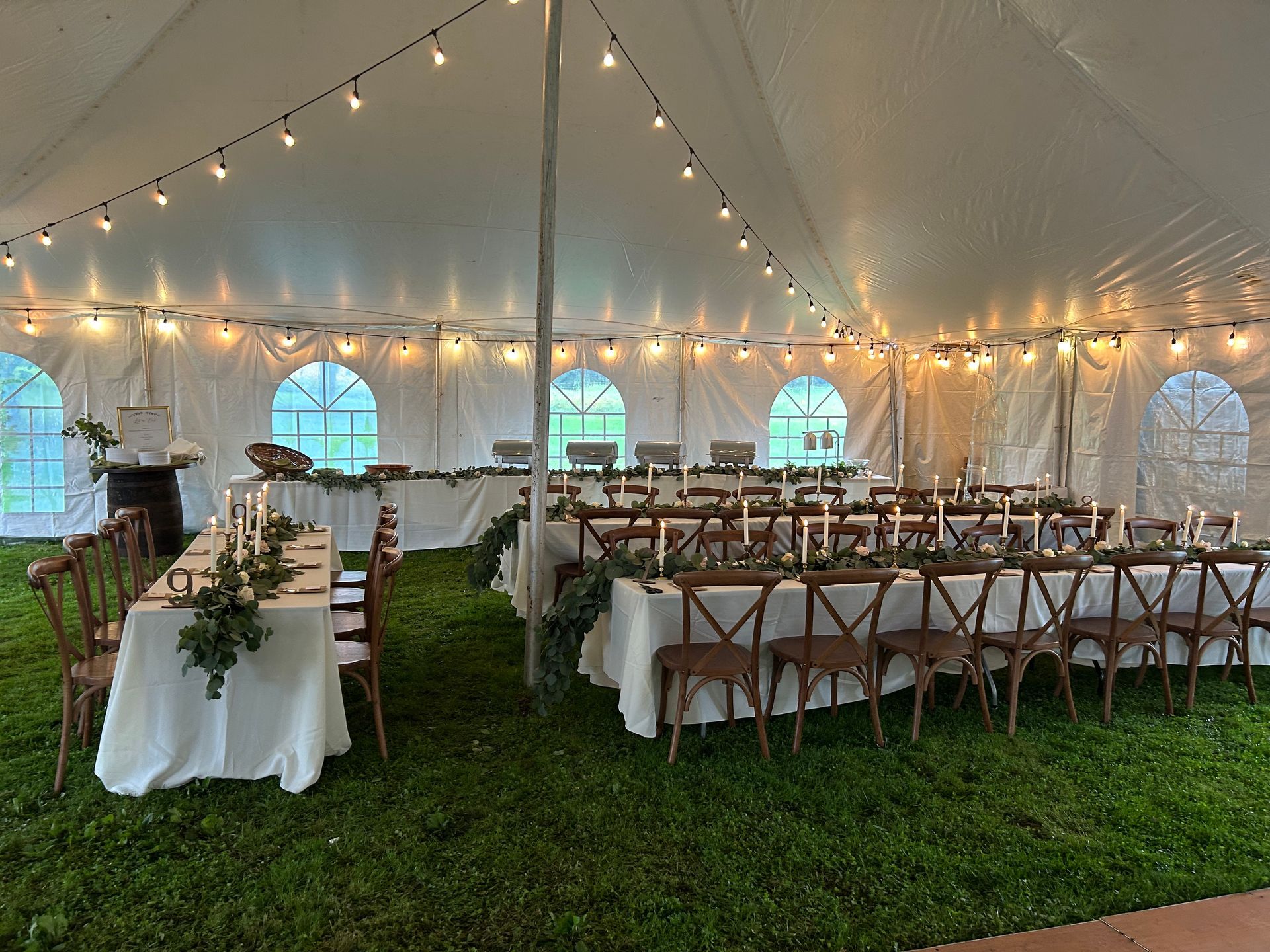 Wedding tent reception with tables, chairs, string lights, and greenery.