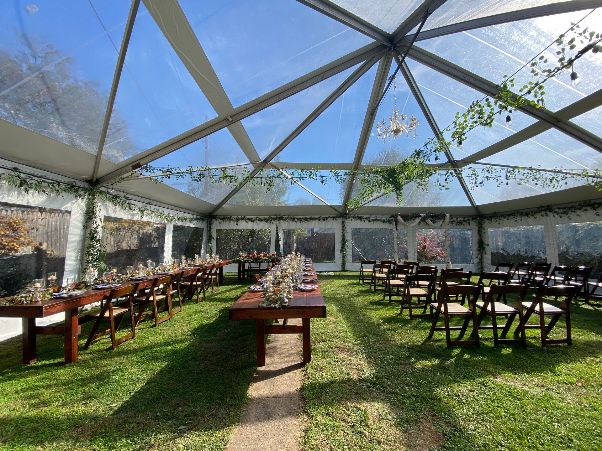 Wedding reception tent with tables set for guests, on green grass. Clear roof and sides.