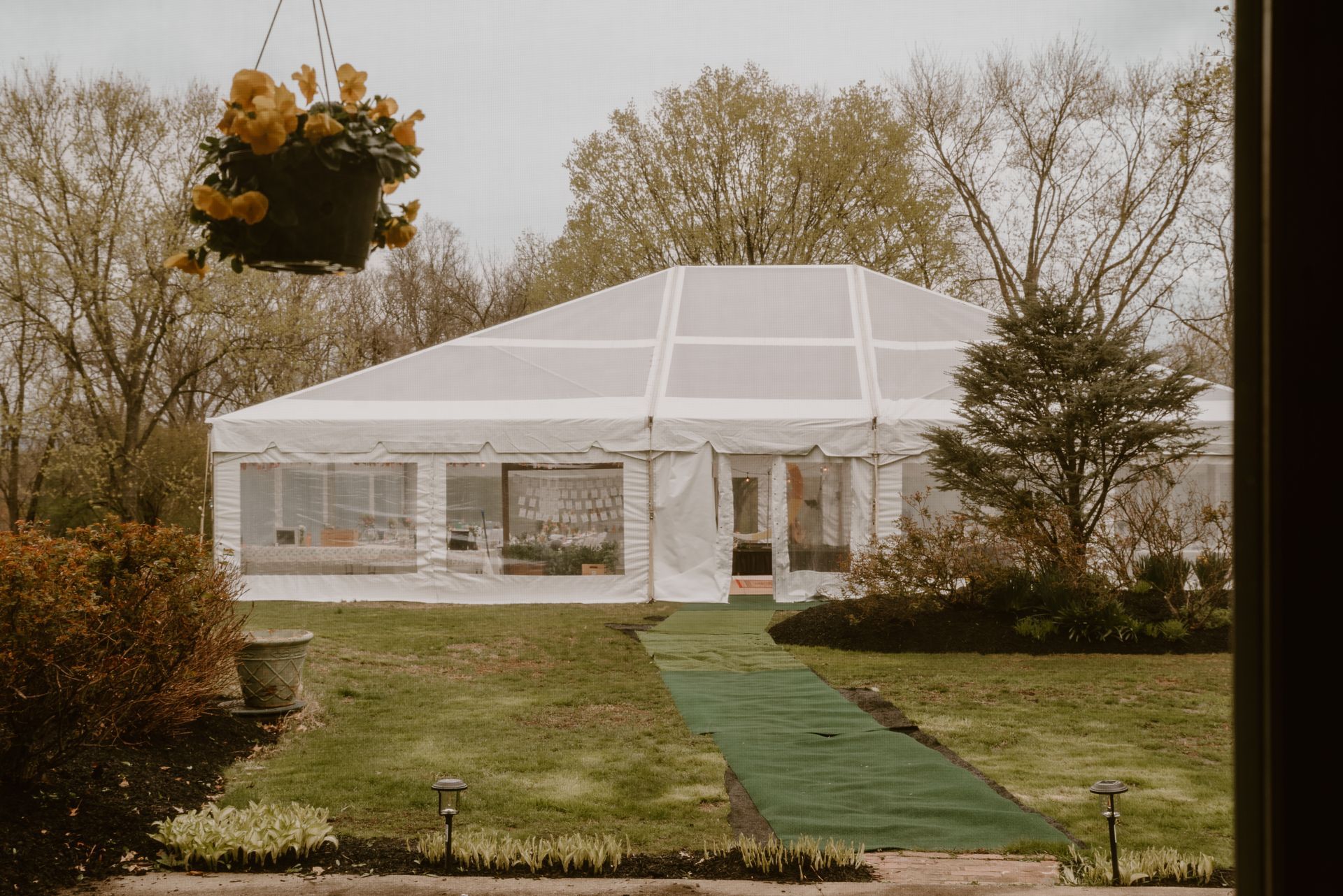 White tent event structure on a green lawn with a hanging flower basket. Trees in the background.
