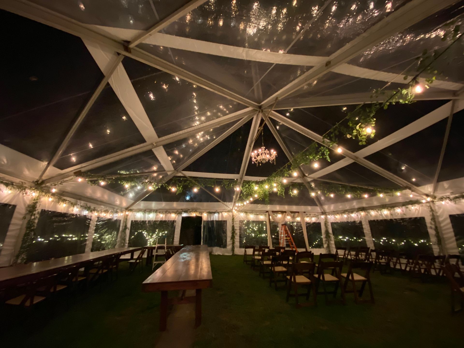 Clear tent interior with string lights, long tables, chairs, and artificial grass.