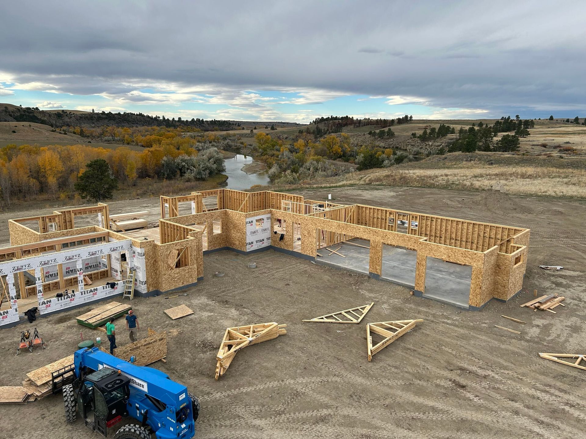 Construction site of a large, wooden-framed house. Workers and a blue tractor. Cloudy day, rural setting.