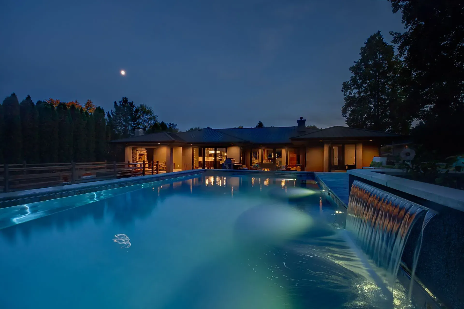 A large swimming pool with a waterfall in front of a house at night.