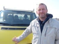 Ein Mann in einer weißen Jacke steht vor einem gelben Lieferwagen.