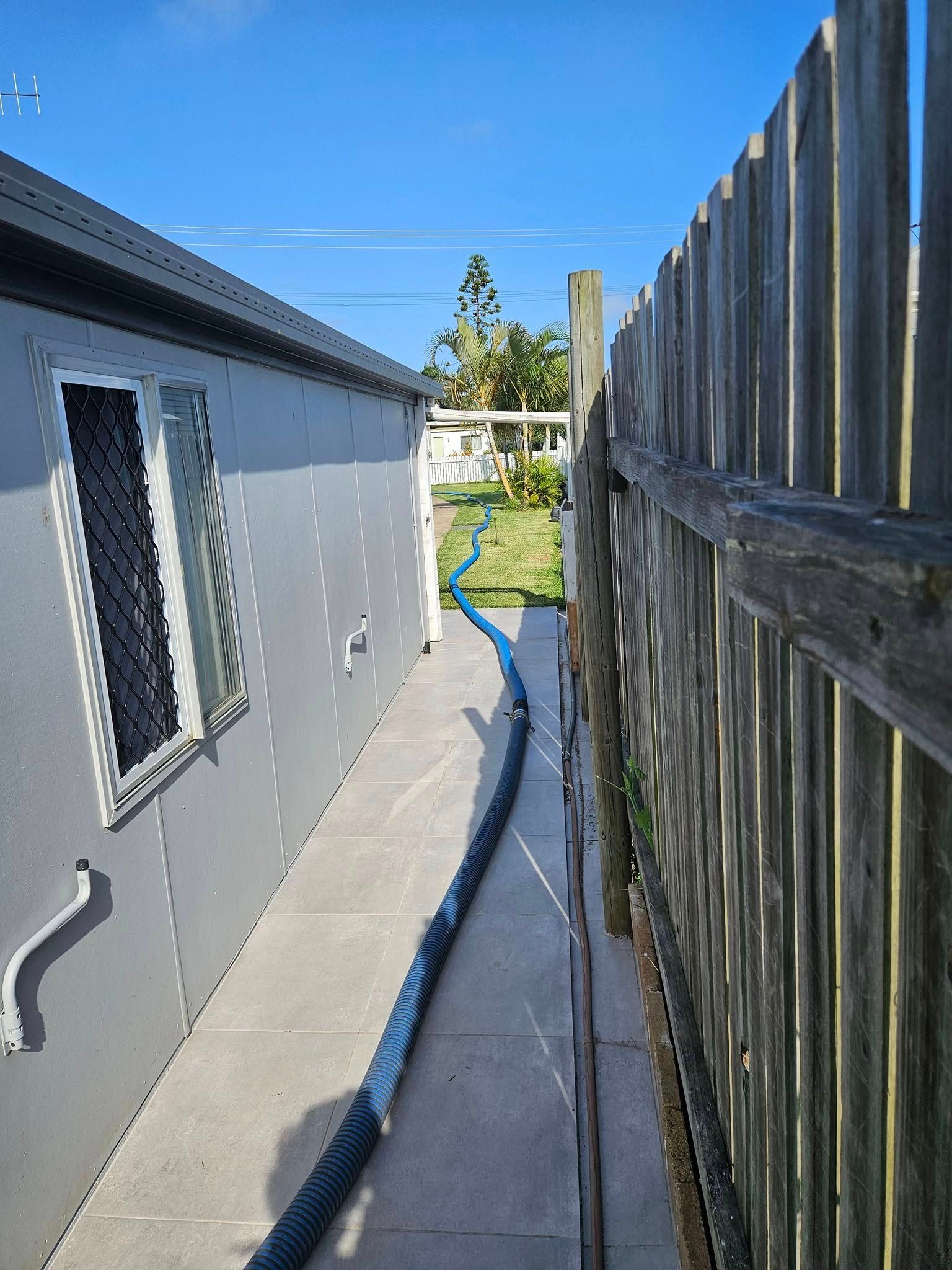 A Blue Hose Runs Between a Gray Building — Paap's Cleansing Services in South Kolan, QLD