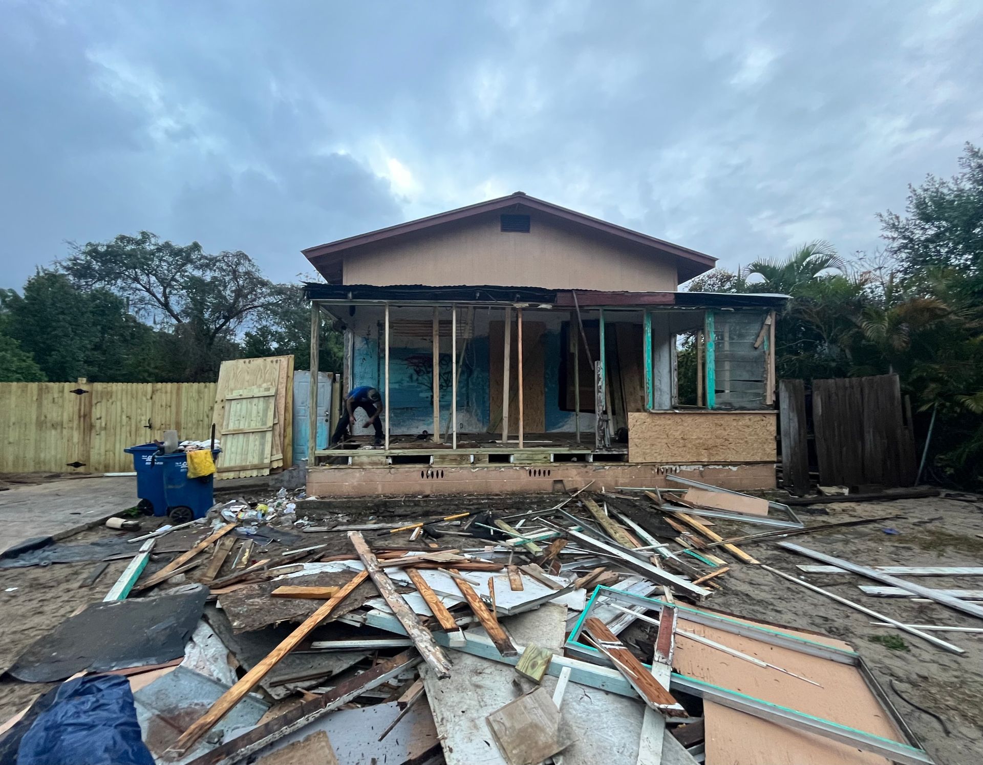 House undergoing renovation, debris in foreground. Construction on porch area, overcast sky.