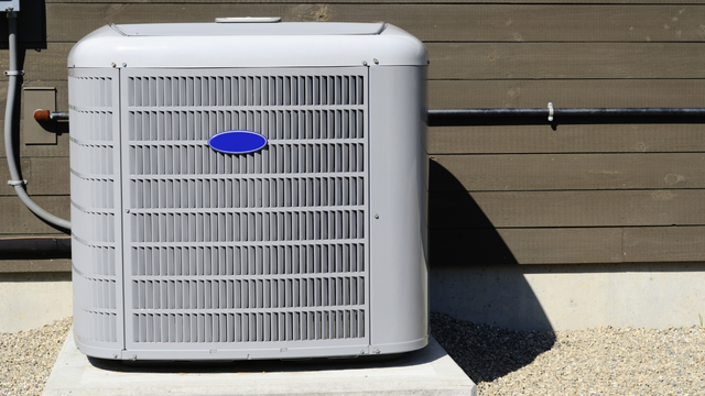 Gray air conditioning unit outside on a concrete base next to a brown wooden wall.