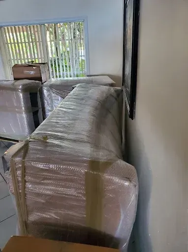 Furniture wrapped in plastic, stacked inside a room, near a window.
