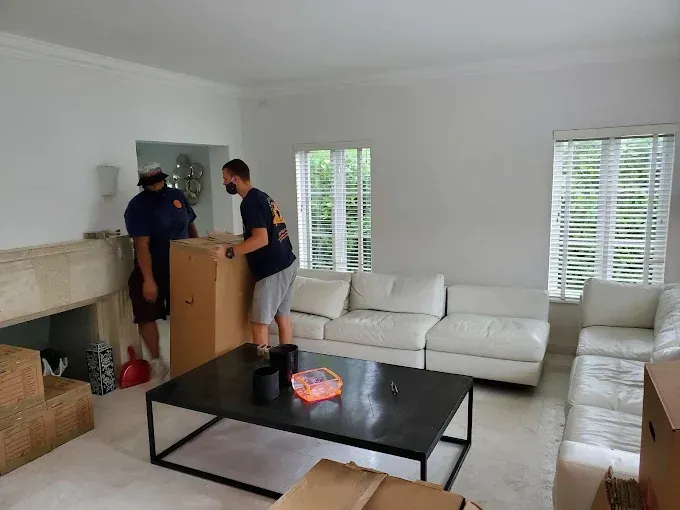 Two movers carrying a large cardboard box in a living room with a white couch, windows, and fireplace.