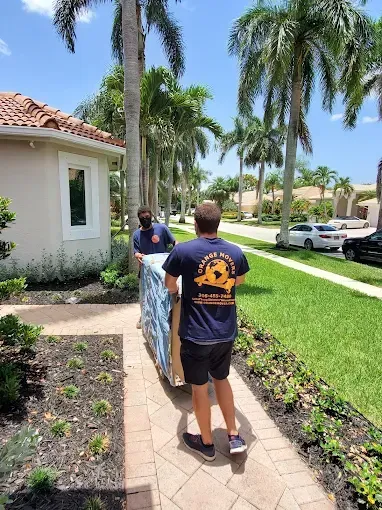 Two movers carrying a large, wrapped item on a sidewalk in front of a house on a sunny day.