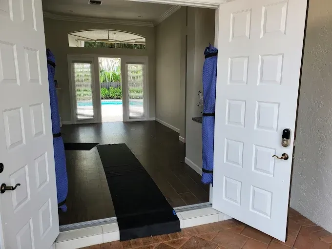 Open double white doors reveal an interior with dark wood floors, blue pool view, and a black mat.