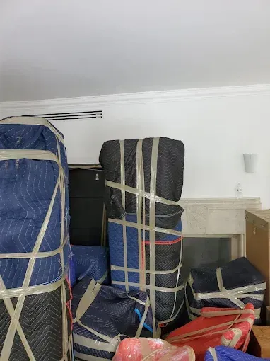 Packed furniture wrapped in blue and black moving blankets, secured with tape, in a room.