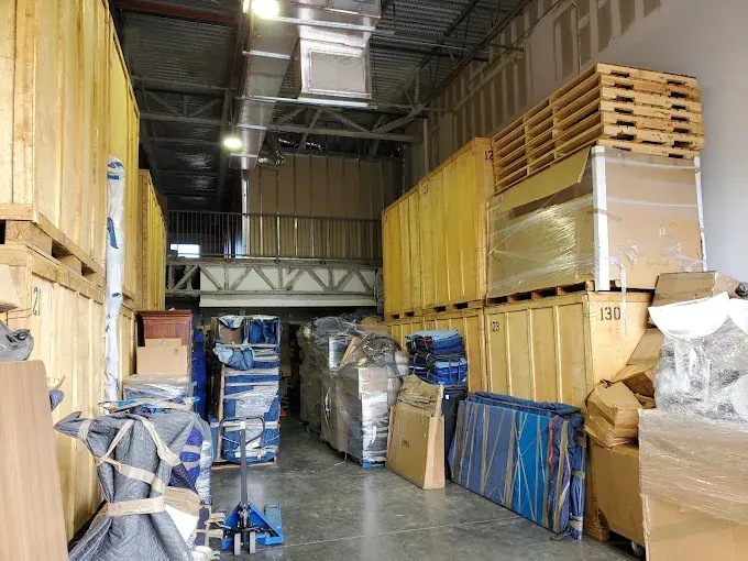 Warehouse interior with stacks of wooden crates, pallets, and wrapped furniture.