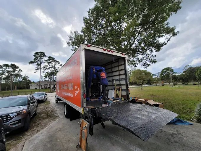 U-Haul moving truck with ramp down. Person loading boxes into the orange truck on a driveway. Cloudy sky, green grass.