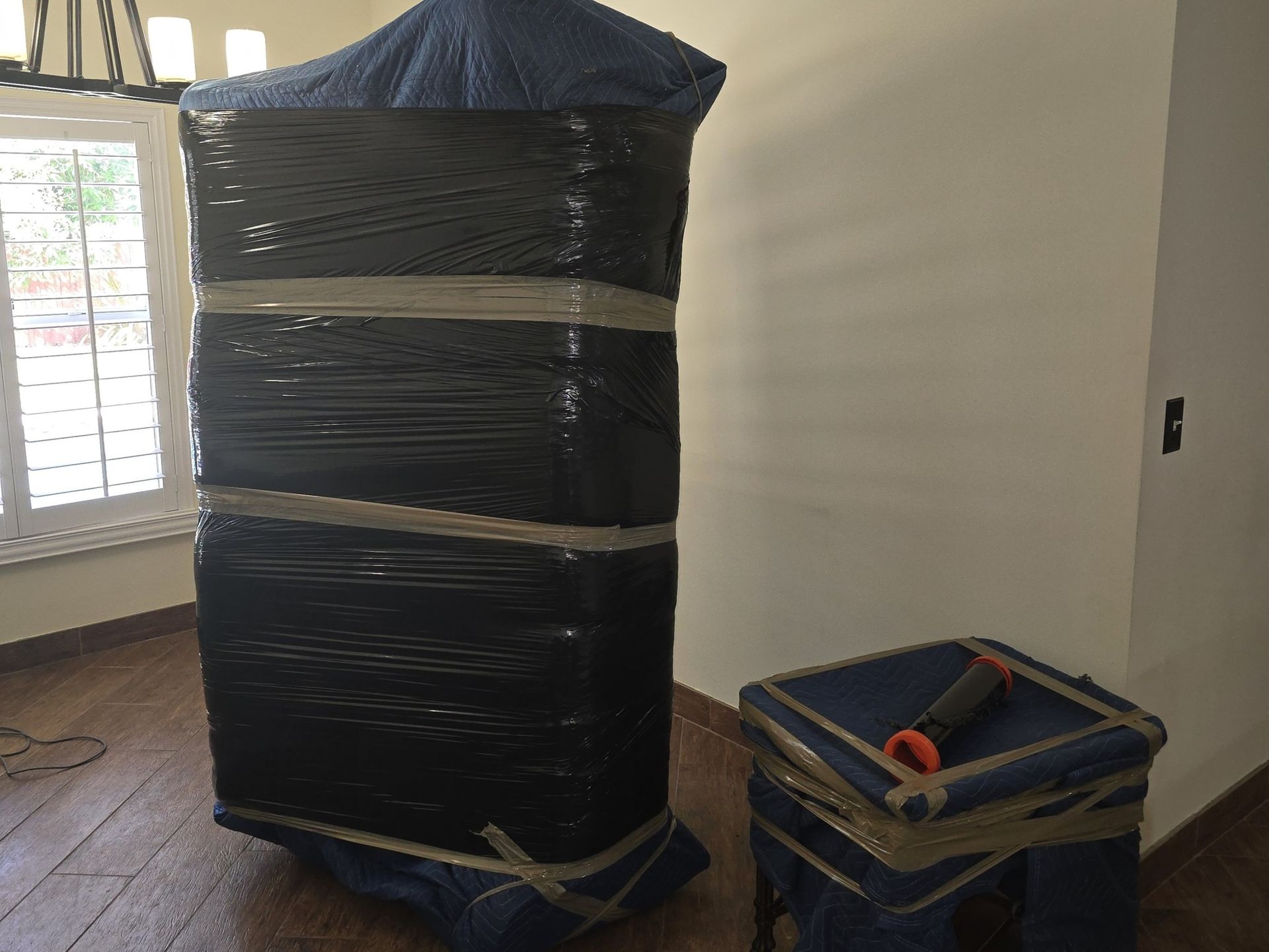 Large furniture wrapped in plastic and tape, prepared for moving. Small, wrapped table nearby. Indoors.