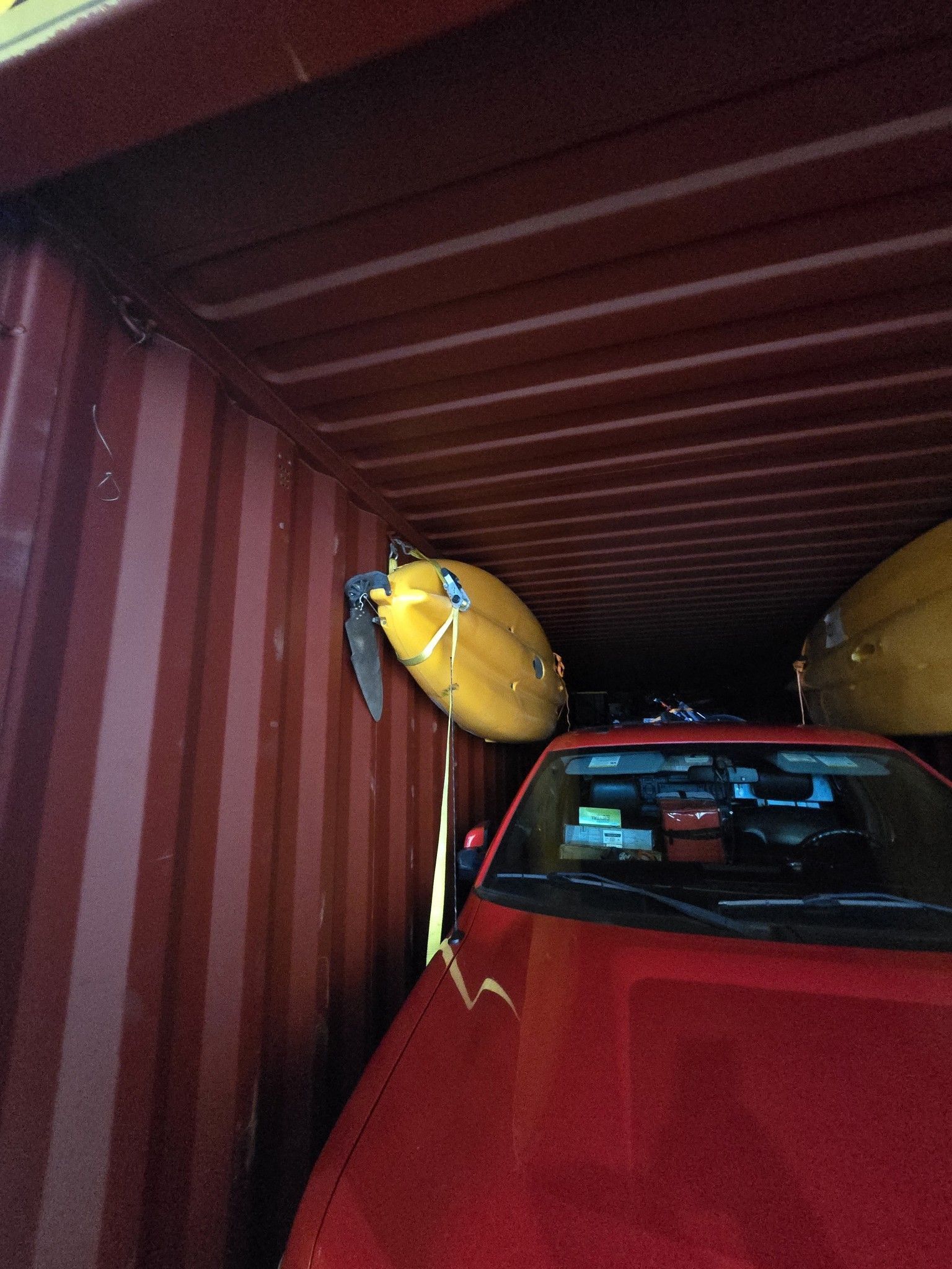 Inside a red shipping container: A red car, a yellow kayak, and a yellow boat.