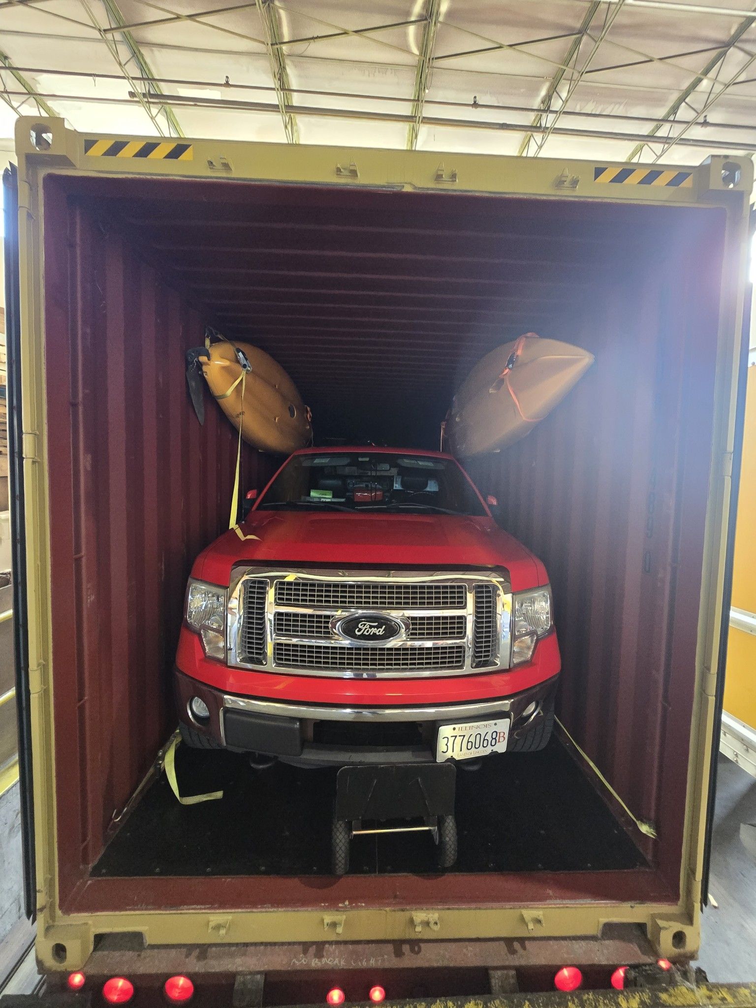 Red pickup truck inside a shipping container, secured with straps. Two kayaks are strapped to the roof.