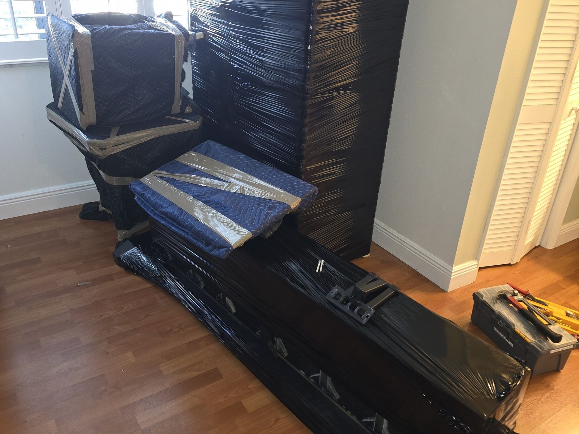 Furniture wrapped in black plastic, ready to move. Includes a chair, table, and other long items near a doorway.