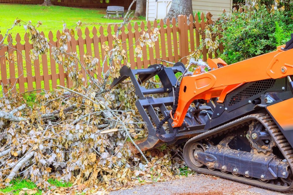 Orange skid steer with grapple collecting debris next to a brown wooden fence.