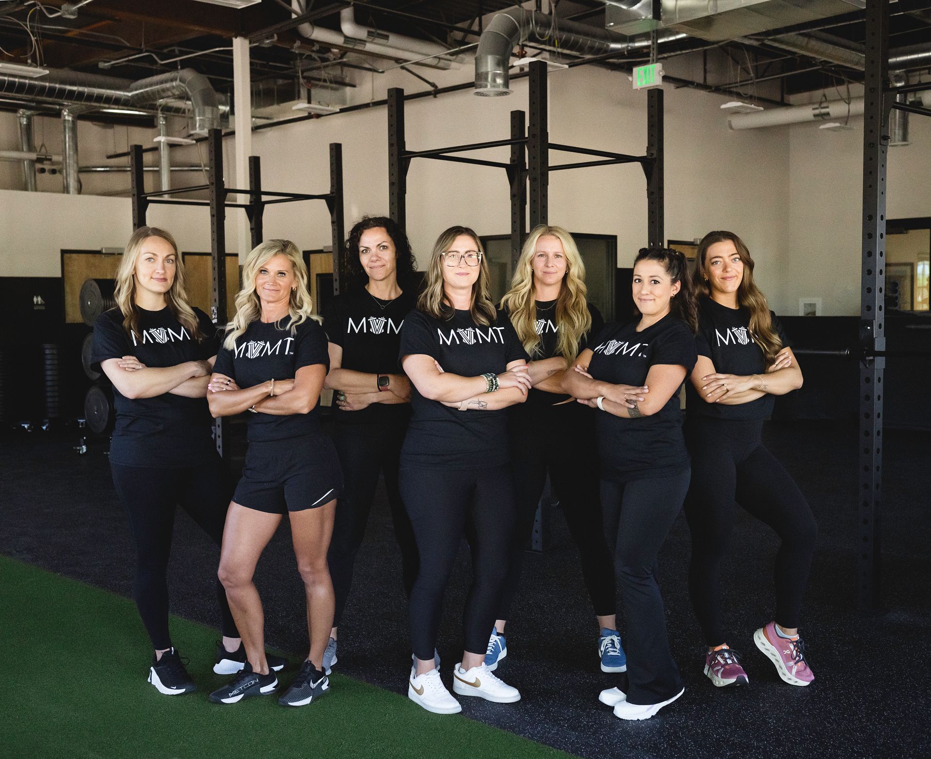 Group of seven people with arms crossed, in a gym. They are wearing black shirts and pants.