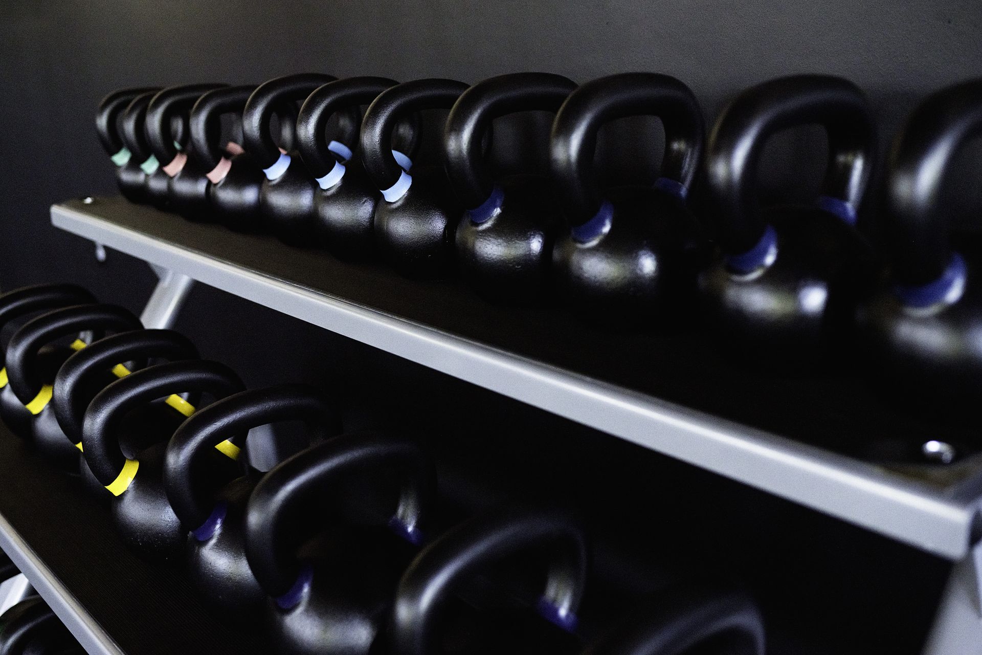 Rows of black kettlebells with color-coded handles on a rack in a gym setting.