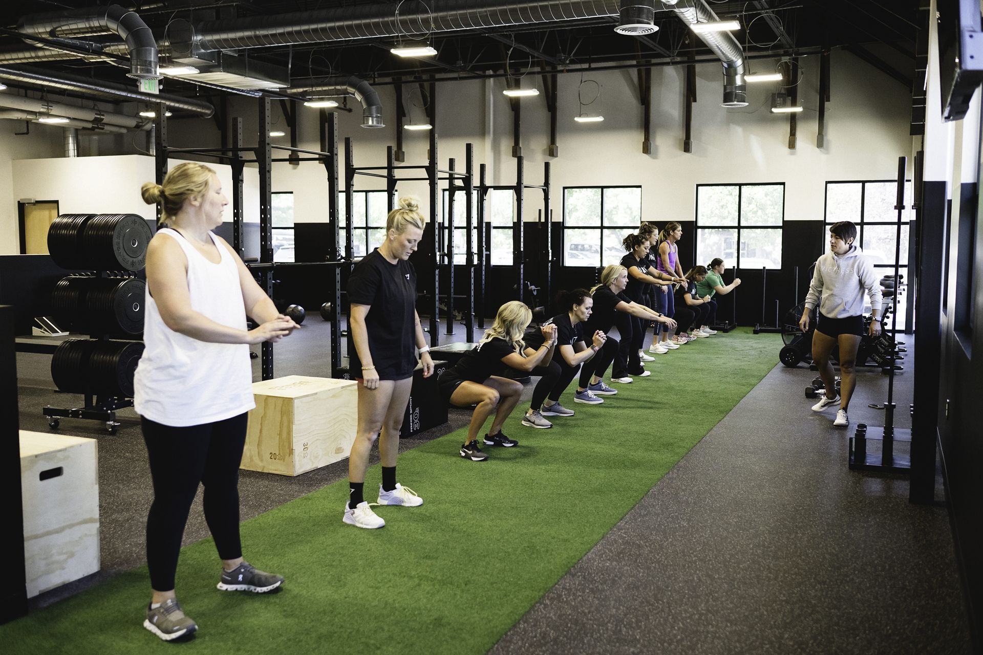 People doing squats in a gym. Trainer observes, others in line, on green turf, near equipment.