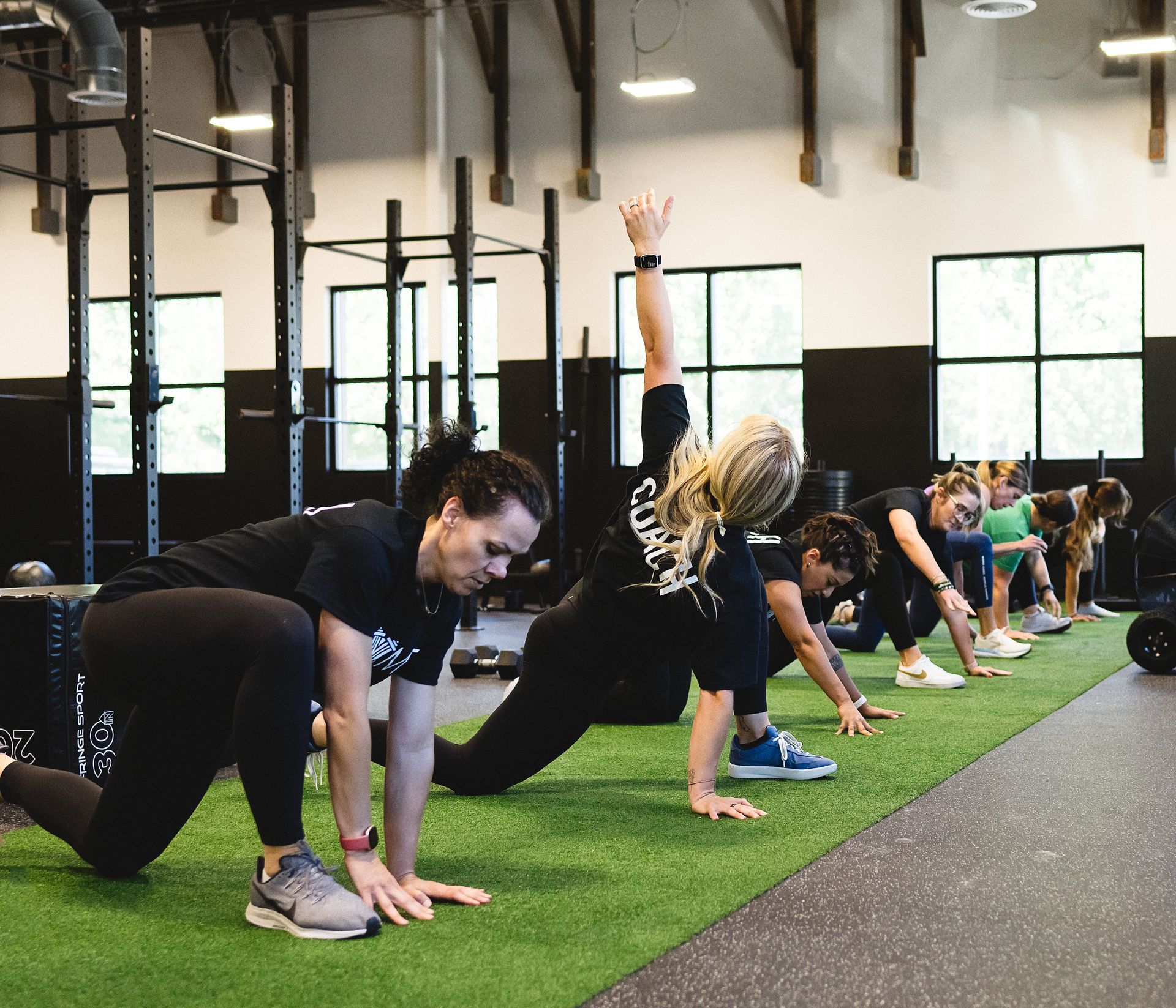 People stretching on green turf in a gym. One woman reaches overhead. Black athletic attire.