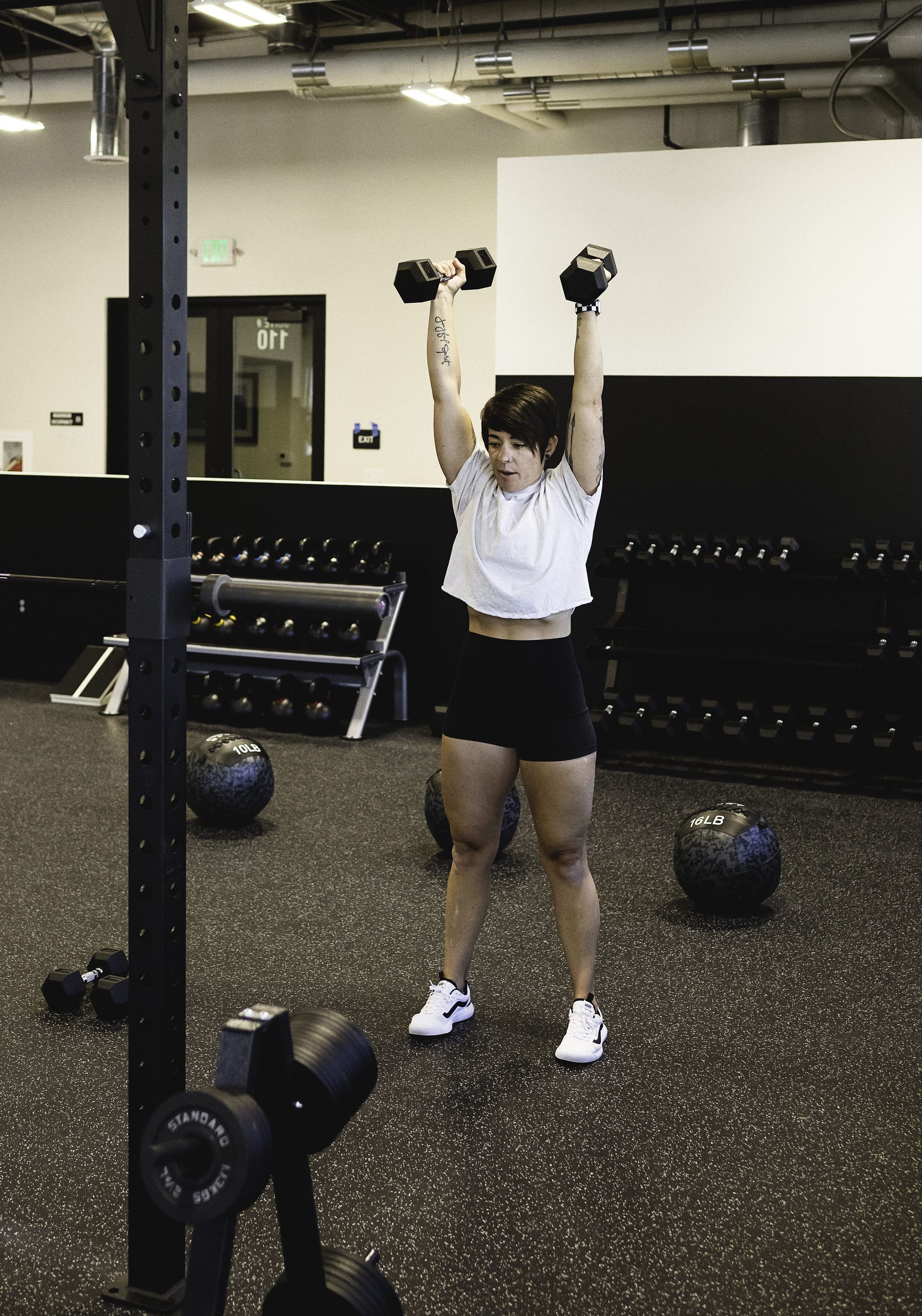Woman in gym lifting dumbbells overhead. Black shorts, white top, and sneakers.