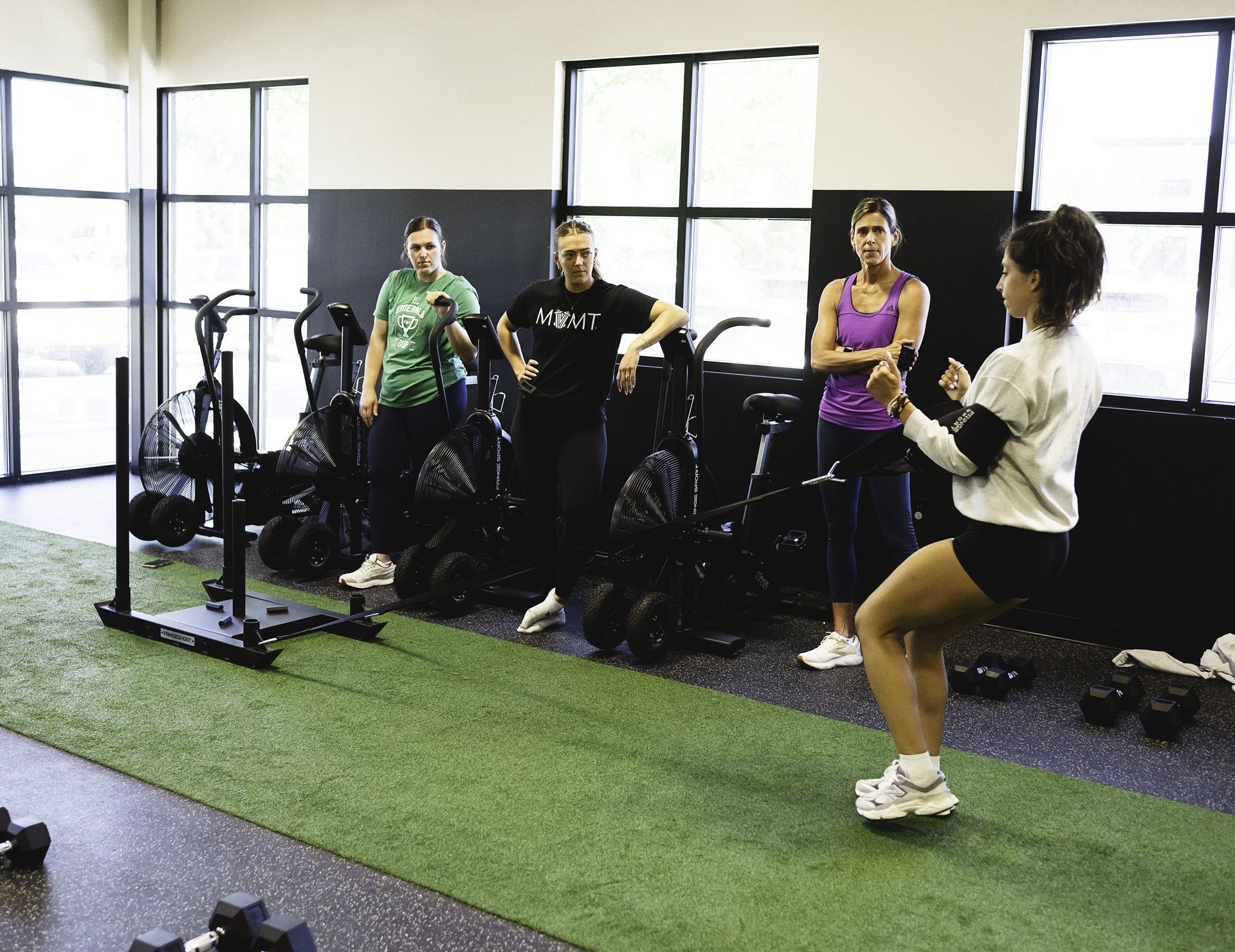 Woman demonstrating a workout; three others watch in a gym. Green turf, exercise equipment, and windows are visible.