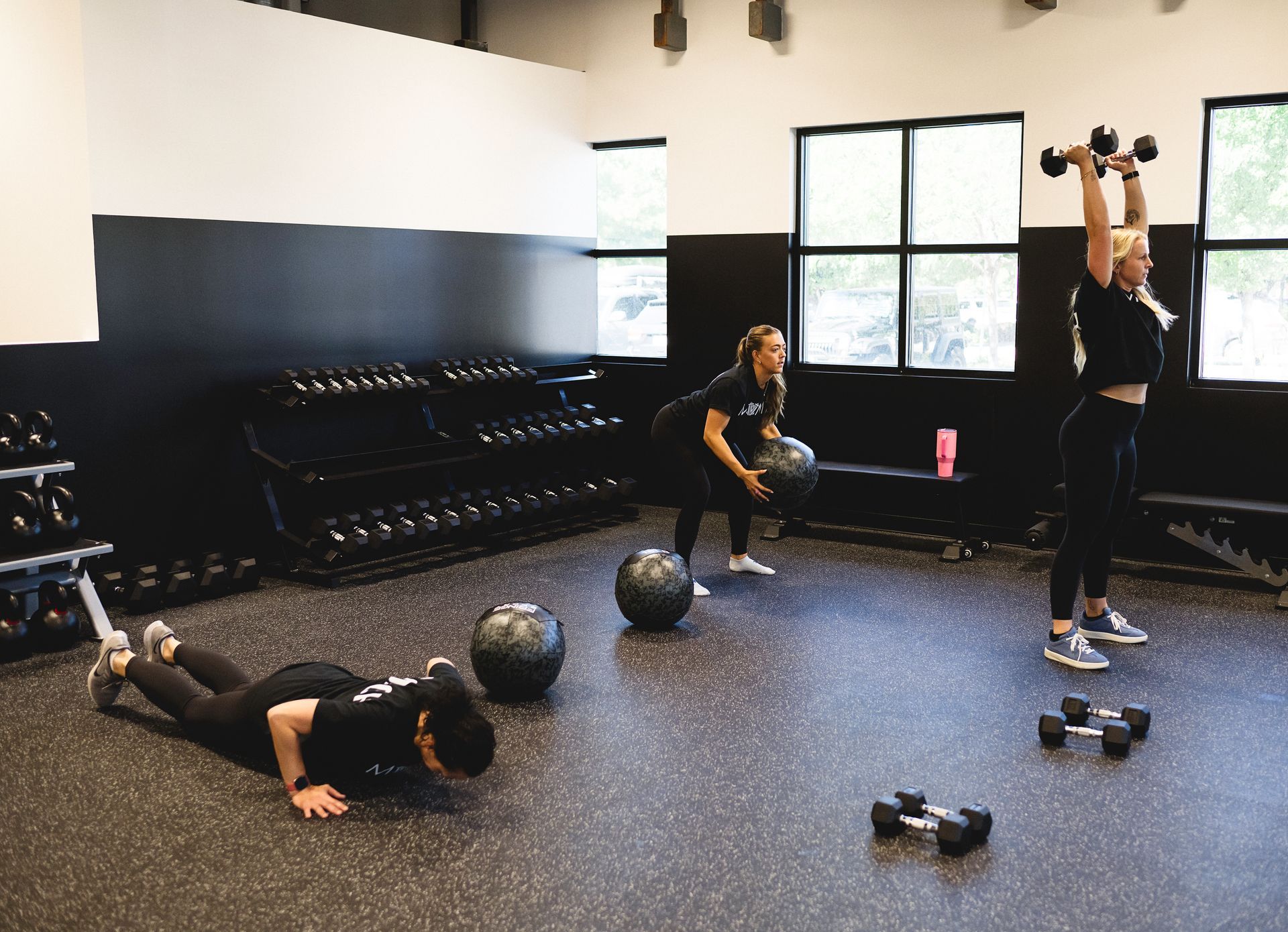 People working out in a gym. One does push-ups, one holds a medicine ball, and one lifts dumbbells overhead.