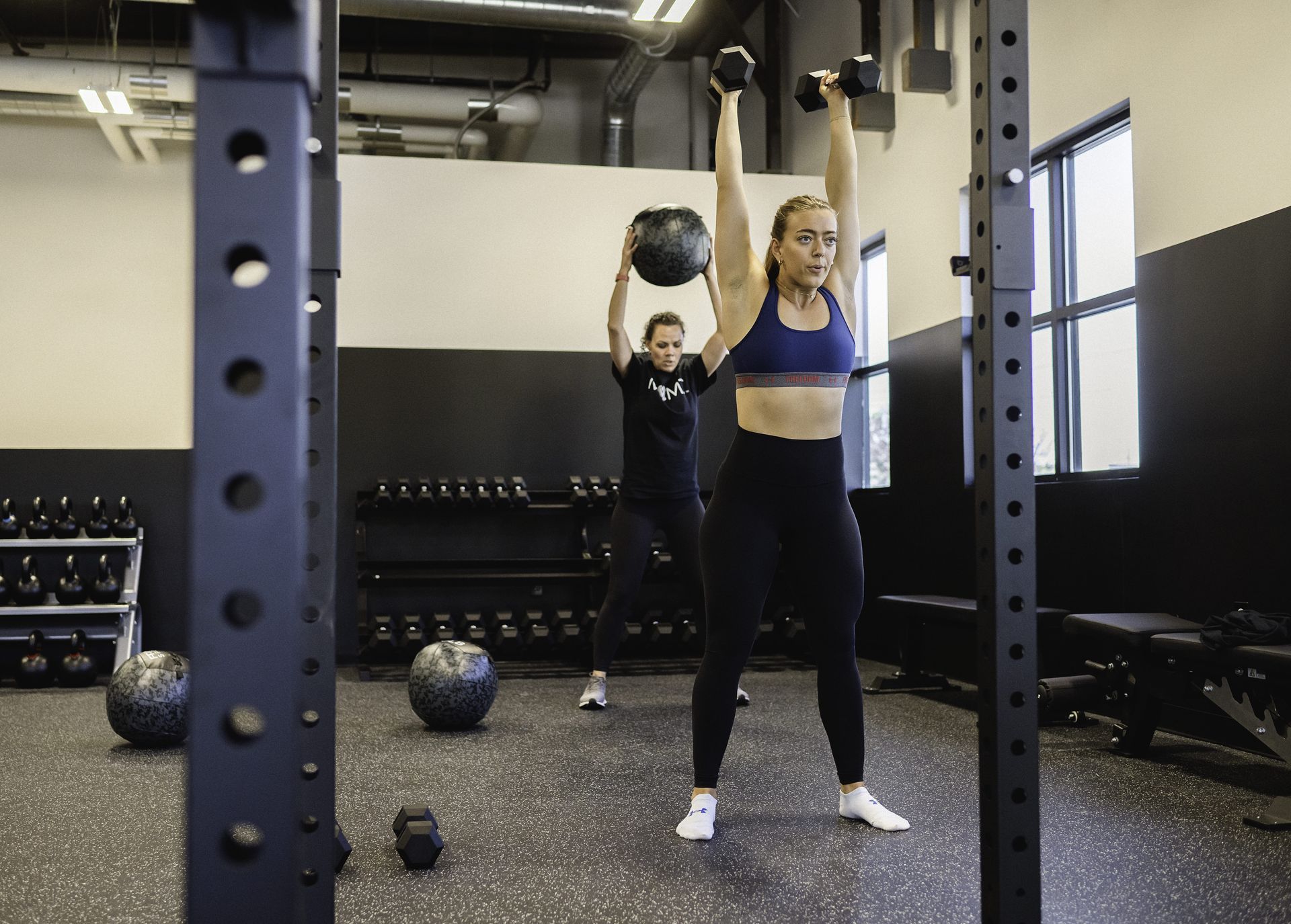 Two people exercising in a gym: One holding dumbbells overhead, the other with a medicine ball.