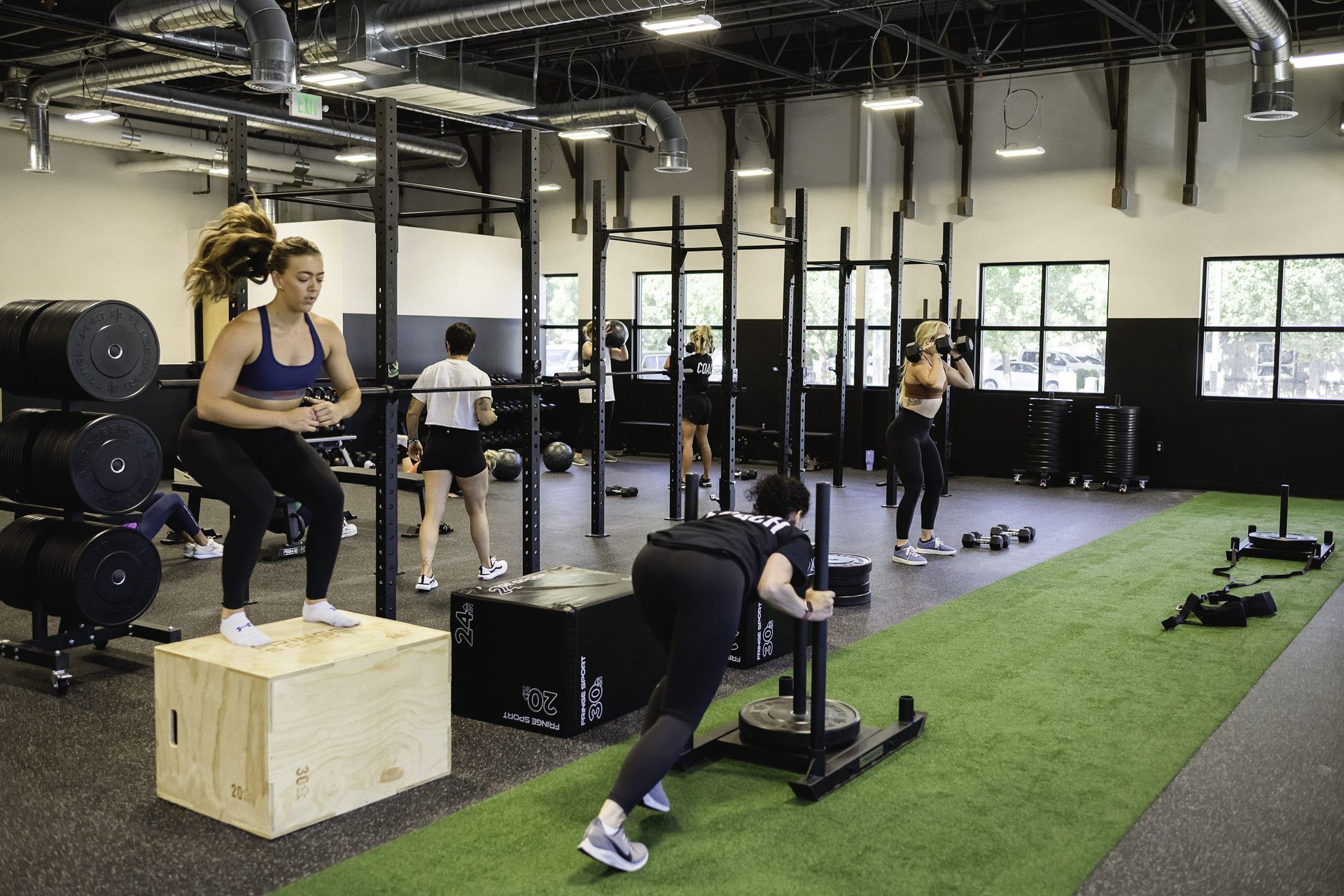 People working out in a gym, one woman jumps on a box, another pushes a sled on turf.