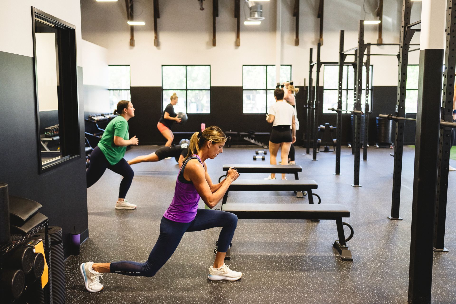 People exercising in a gym, doing lunges, with weights and exercise benches present.