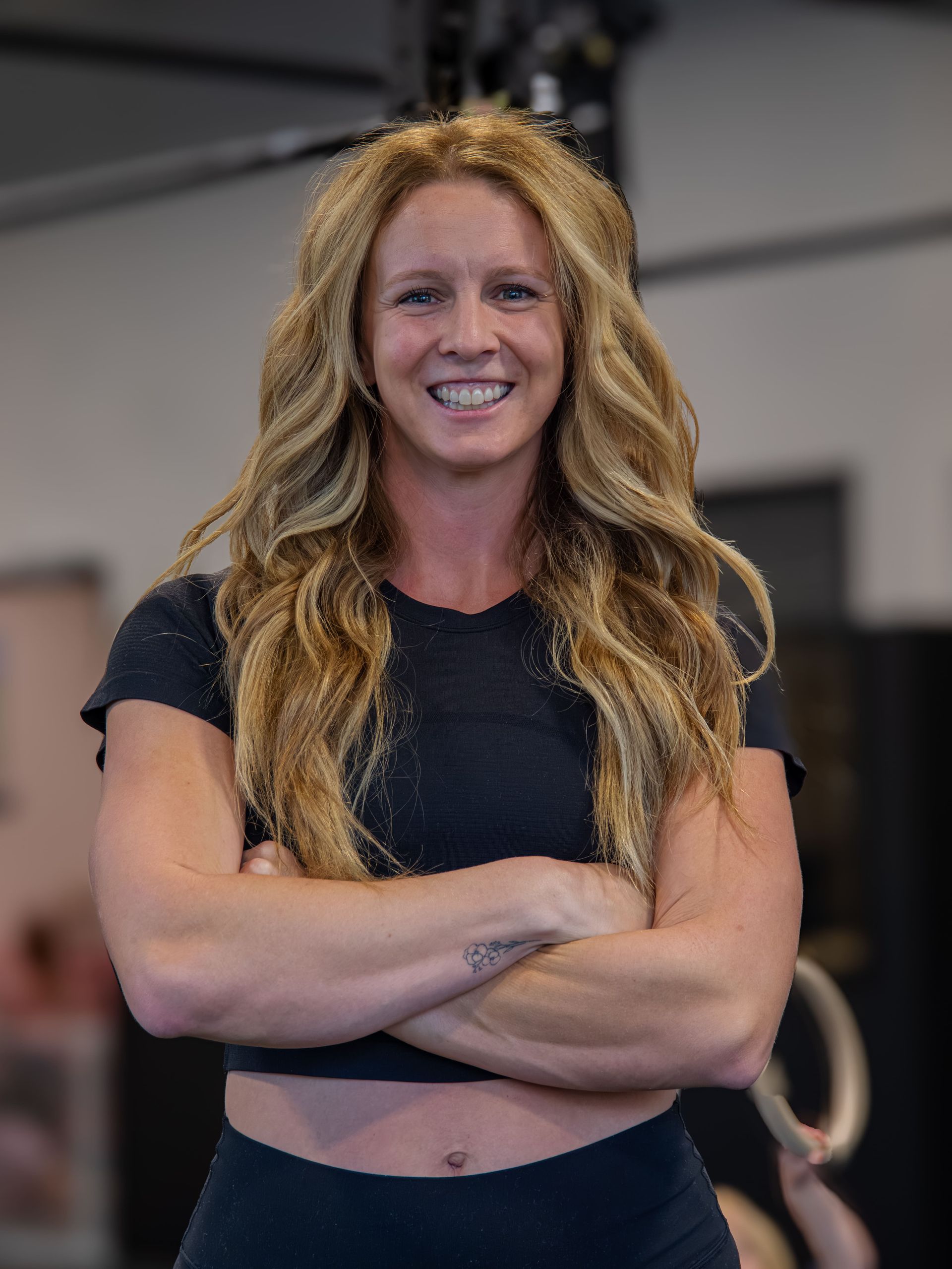 Woman with wavy blonde hair, arms crossed, smiling in a gym setting.