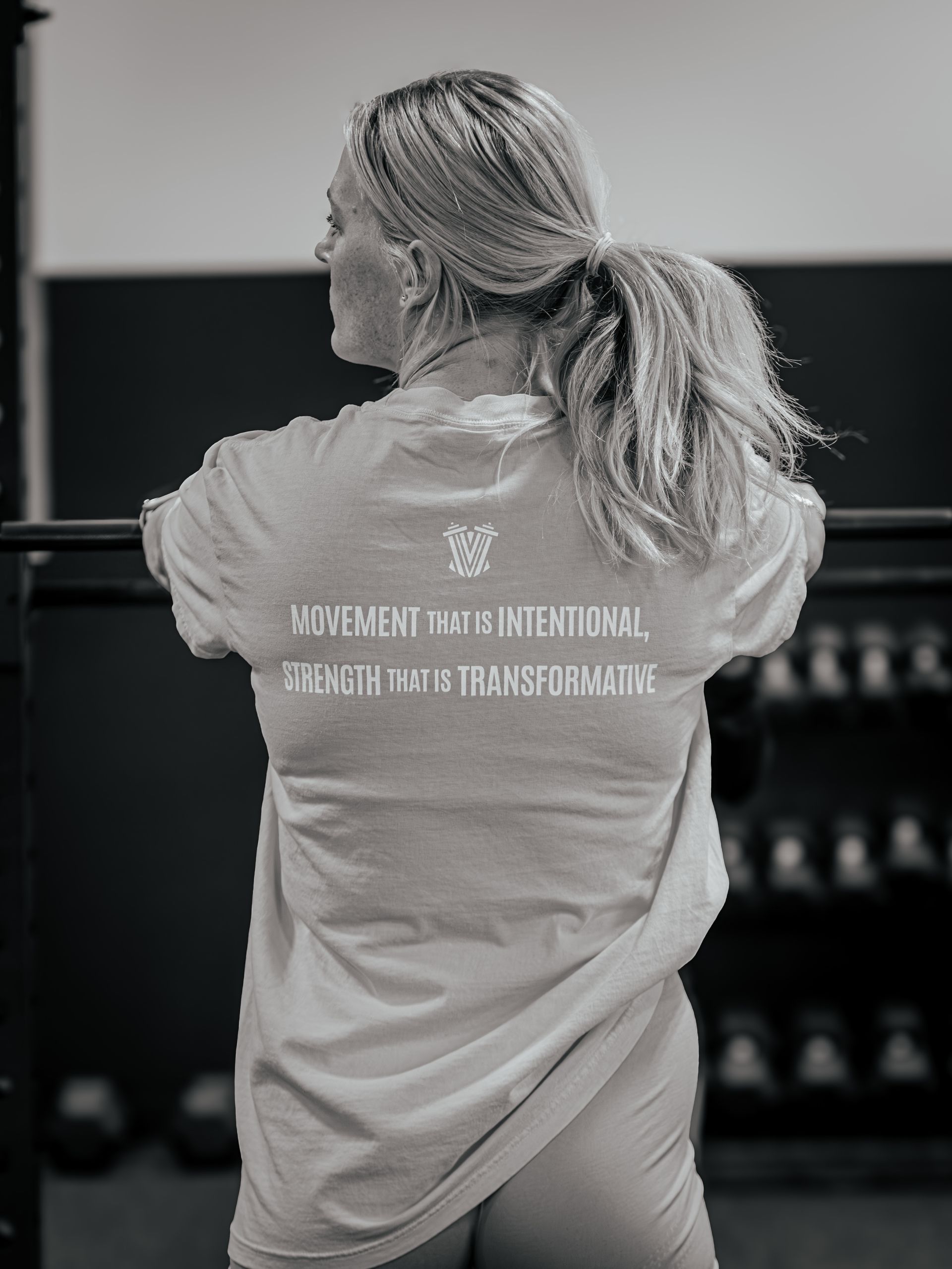 Woman in a t-shirt leaning on a barbell, back to the camera, in a gym. Text on shirt: 