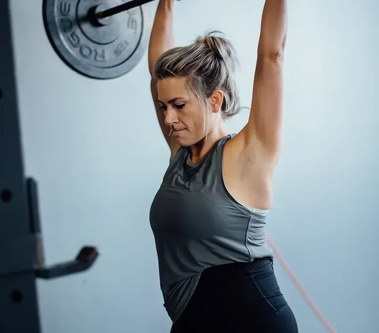 Woman lifting barbell overhead in a gym. She wears black leggings and gray tank top, focused expression.