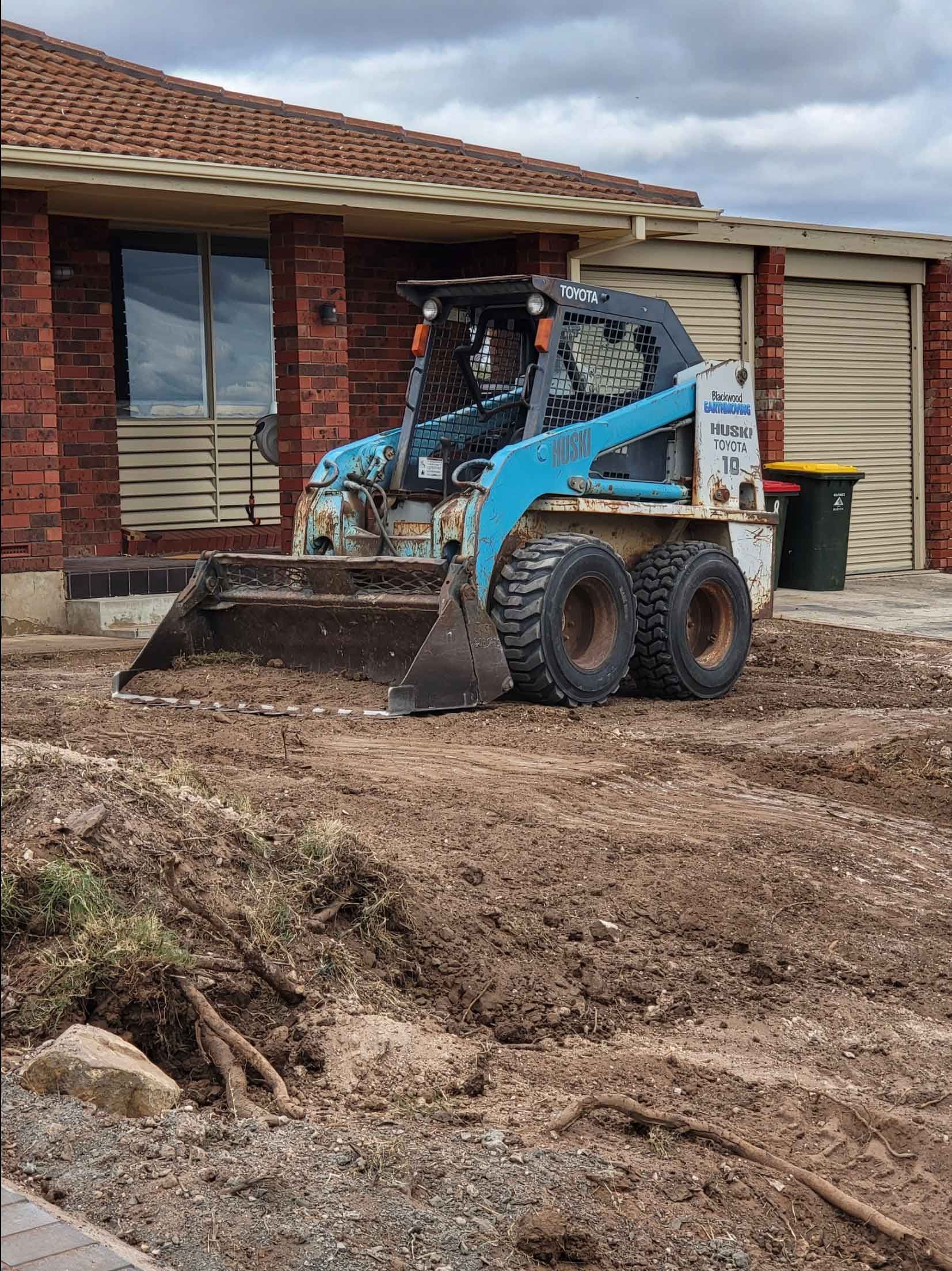 A Bulldozer Is Moving Dirt In Front Of A Brick House – Blackwood SA - Blackwood Earthmoving