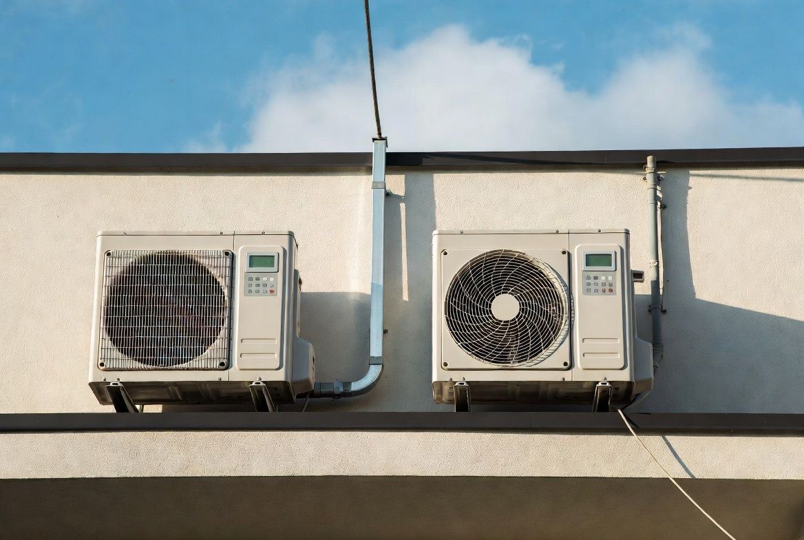 Two wall-mounted air conditioners beneath a roofline against a blue sky.