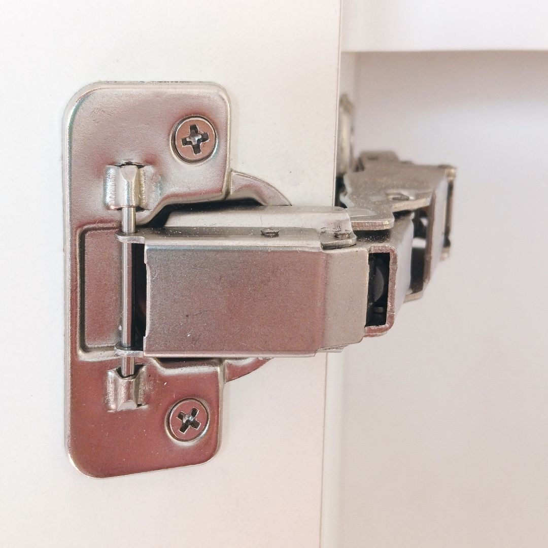 Primer plano de una bisagra plateada para armario montada en un marco de puerta blanco.