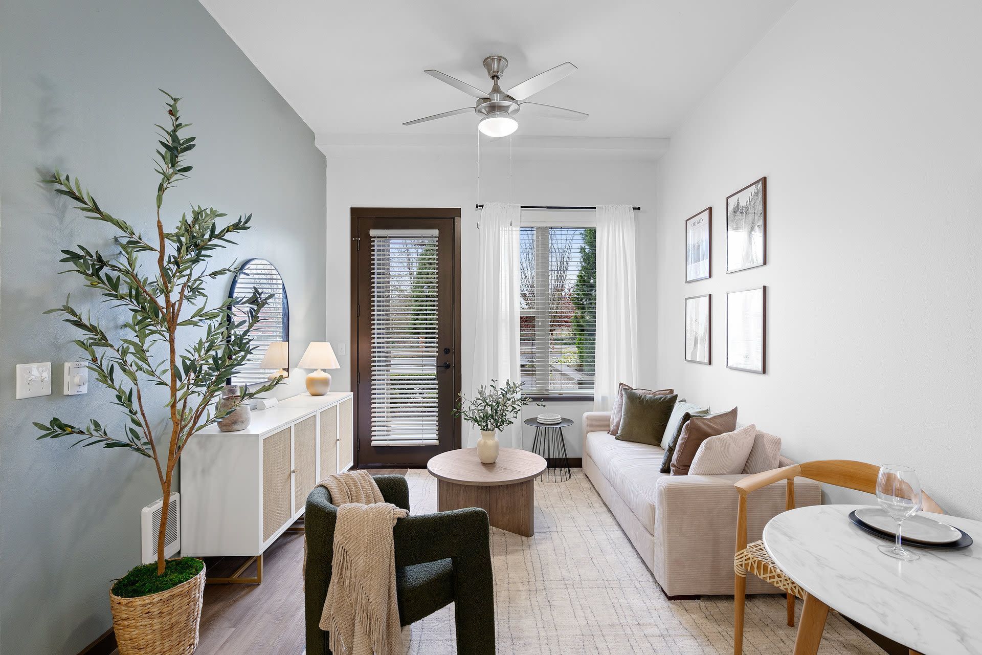 Bright living room with beige sofa, round coffee table, ceiling fan, and glass door with blinds.
