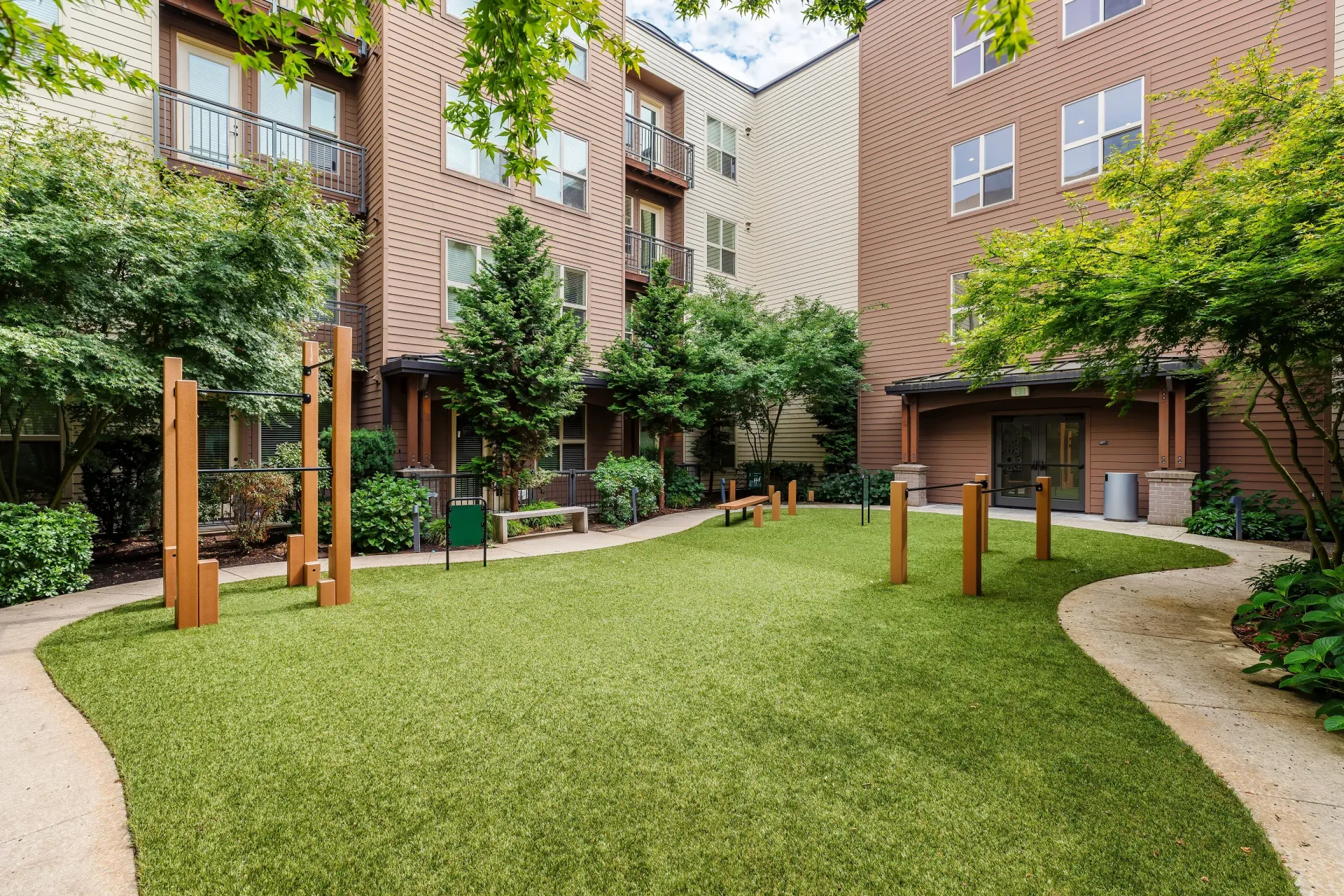 Outdoor apartment courtyard with green lawn, trees, benches, and exercise posts.