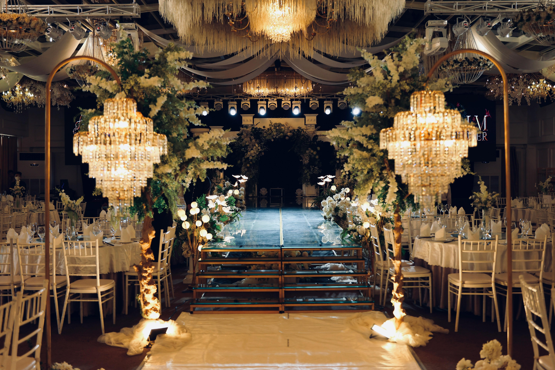 Elegant chandelier-lit wedding aisle with gold chairs and floral archway in a dark, ornate hall